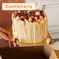 Pessoa segurando bolo decorado com frutas vermelhas e raspas de chocolate branco, sendo colocado em embalagem de confeitaria.