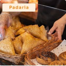 Atendente organizando salgados em uma cesta dentro de vitrine de padaria, com destaque para a variedade de produtos assados.